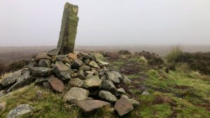 A tall, weathered boundary stone stands upright on top of Cock Howe, rising from a heaped cairn of grey and rust-coloured rocks. A faint carved mark is visible near the top of the stone. Behind it, the moor fades into thick mist, with tussocks of grass and low gorse scattered across the mound and the ground beyond.