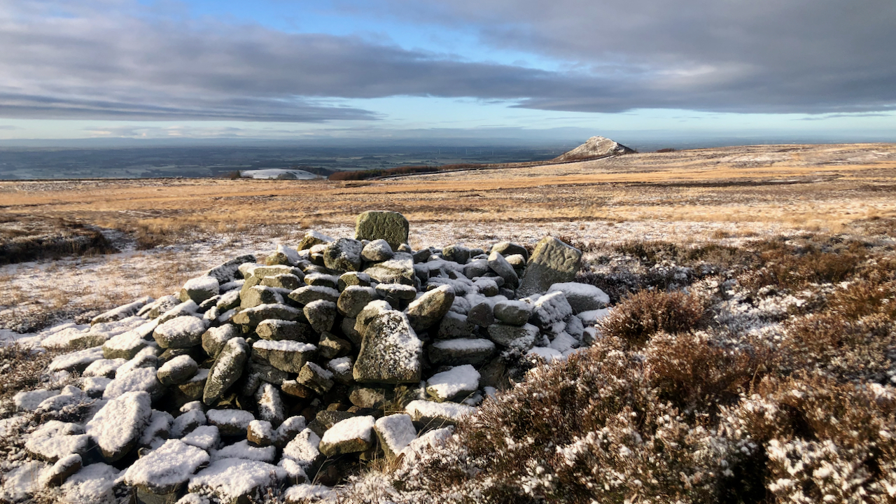 View of Great Ayton Moor with a snow encrusted cairn in the foreground and Roseberry Topping in the distance.