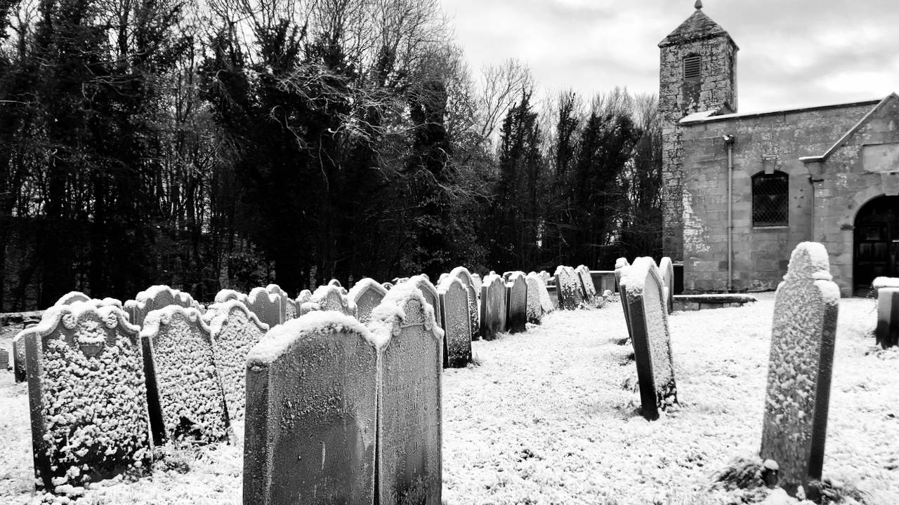 A black and white photo of snow covered gravestones in the churchyard of St. Andrew's Church, Ingleby Greenhow.