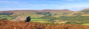 A panoramic landscape photo from a hilltop. In the foreground, there is a large, weathered rock formation. In the distance, rolling hills covered in green fields and patches of trees stretch towards a clear blue sky.