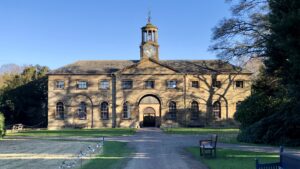 The stables at Ormesby Hall, a large historic building with a clock tower. The stables feature a symmetrical design with a central entrance and a row of arched windows on either side. The clock tower rises above the centre of the building, adding a distinctive feature. The building is made of stone and has a slate roof. The surrounding area is well-maintained, with a paved path leading up to the building and benches placed nearby.