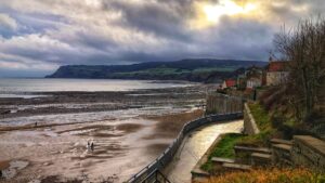 A scenic coastal town nestled against a cliffside. The sun shines through the clouds, casting a warm glow on the beach and the colourful houses. A few folks stroll along the beach, enjoying the beautiful scenery. It is low tide.