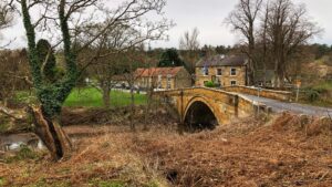 A rustic stone bridge with a single arch crosses a river. The bridge is made of large, ashlar stones and has a road crossing over it. Along the riverbank, there is dry bracken and a few trees. A large ivy-covered with a hollowed out inner dominates the foreground. In the distance, a village with a pub and several other buildings is visible. The trees in the background are bare and the sky is overcast.