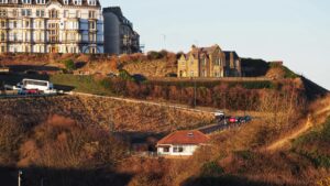 A two-story stone house sits on a steep cliff below the coastal town of Salt urn-by-the-Sea. Cars and a bus navigate along the steep road up the cliff. Top left are terraces of four-story dwellings. There is a red-roofed commercial property lower down the cliff.