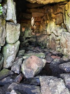 A close-up outdoor shot shows the interior of the stone structure of an horizontal mill. The structure is built from large, rough-hewn stones, creating a dark, enclosed space. Light filters in from the entrance, illuminating the uneven stone walls and the rocky ground inside. Some of the stones are covered in moss or lichen, suggesting a damp environment. The overall impression is of an ancient, weathered structure.