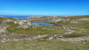 An eye-level, wide shot shows a somewhat barren, rocky landscape dotted with patches of green grass and two small, still pools of dark blue water under a clear, bright blue sky. The foreground features uneven terrain with exposed light-coloured rocks and areas of short, green grass. A shallow, dry streambed winds through this area. Before and to the left of the pools, a ruined medieval chapel sits on a small knoll. Further back, the two small, circular pools of water reflect the clear sky. Around the pools, the landscape is rockier. In the far distance, a strip of deep blue ocean meets the horizon under the expansive blue sky. The overall scene conveys a sense of remote, windswept moorland on a sunny day.