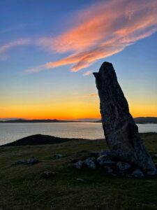 A striking silhouette of MacLeod's Stone stands against a vibrant sunset. The sky is a gradient of deep blue at the top, transitioning to fiery orange and yellow near the horizon, with wisps of pink and orange clouds stretching across. The standing stone, dark and imposing, is angled slightly, with its base surrounded by smaller, shadowed rocks. The sea reflects the fading light in the background, with the dark outlines of distant land visible across it. The immediate foreground is a grassy slope, mostly in shadow.