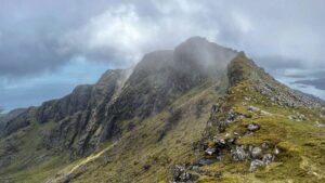 An expansive, eye-level shot capturing the mountain ridge of Beinn Mhòr on South Uist on a cloudy day. The ridge, covered in green grass and scattered rocks, stretches from the foreground towards the left, eventually meeting a towering summit peak shrouded in mist. To the right, another misty subsidiary peak is visible, and the sea and lochs can be seen in the distance on both sides of the ridge.