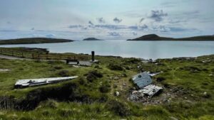 In an expansive outdoor shot, a desolate landscape under a cloudy sky shows parts of a plane crash. In the immediate foreground on the right, large, twisted pieces of metal from the wreckage are strewn across a grassy hillside. To the left, further up the hill, another significant piece of the plane lies flat on the ground. A narrow, gravelly path with a reddish fence runs from the left down to a monument. In the middle ground, a calm, light blue loch stretches out, reflecting the sky. In the distance, several dark, rolling hills rise from the water.