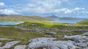 A high view capturing the vast, green, hilly landscape of the Outer Hebrides. In the foreground, large, grey, flat rocks are scattered amidst patches of grass. The midground features rolling green hills with visible sheep paths, leading down to a cluster of small white houses with dark roofs. Beyond the houses, a calm blue loch, dotted with several small boats, extends towards the horizon, where more hills rise. To the left, a turquoise bay with a sandy beach glistens. The sky is partially cloudy with patches of blue.