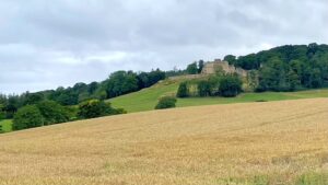 A distant view of Kirby Knowle Castle, a stately home, situated on a hill amidst lush green trees under a cloudy sky. In the foreground, a golden field of crops fills the lower half of the image. The grand, light-coloured castle, with its distinctive roof, is visible in the upper mid-ground, surrounded by dense woodland that extends up the hillside and beyond.