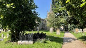 A low-angle, sunlit view shows a well-maintained path winding through a verdant churchyard in the foreground, leading towards a historic stone church with a tall, pointed spire in the background. Lush green trees and grass frame the scene, with various gravestones scattered throughout the cemetery. A prominent, ornate black metal fence encloses a grave in the immediate foreground, while the sunlight casts shadows, highlighting the textures of the stone and foliage.