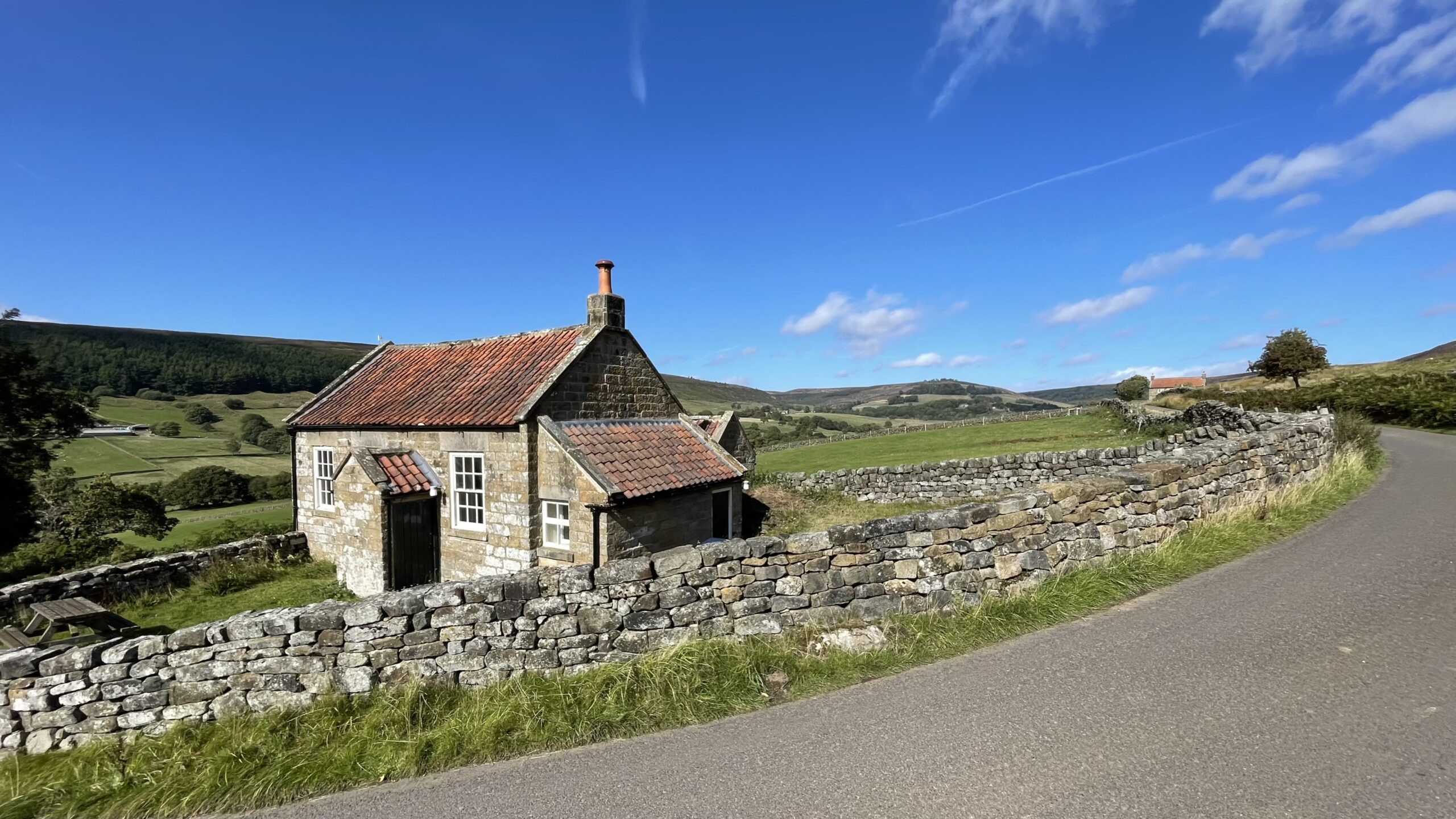 A small, single-story stone building, a former schoolhouse now serving as a community centre, stands next to a grassy field. A low, curved dry-stone wall separates the property from a winding road. In the background, rolling green hills rise under a clear blue sky.