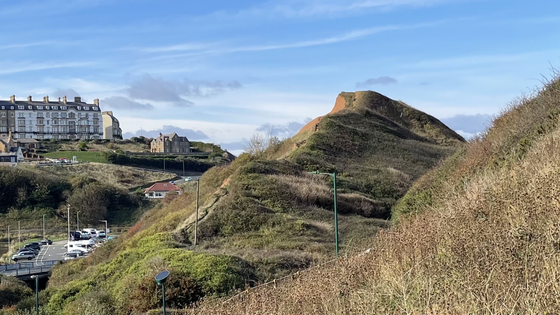 An eye-level view of Cat Nab, a prominent, steep-sided, and roughly conical hill covered in scrubby brown and green vegetation, dominating the right side of the frame. In the distance on the left, at the top of a steep incline, sits a large, multi-story Victorian terrace building with a light-coloured facade. Below the hill and the distant buildings, there is a paved car park visible to the left, with several white vehicles. The foreground is filled with dry, overgrown grasses and shrubs. The sky above is bright blue with scattered white clouds. The entire scene captures the distinct coastal topography of Saltburn-by-the-Sea, showing the transition from the old settlement and beach area (at the base) to the Victorian town on the higher ground.