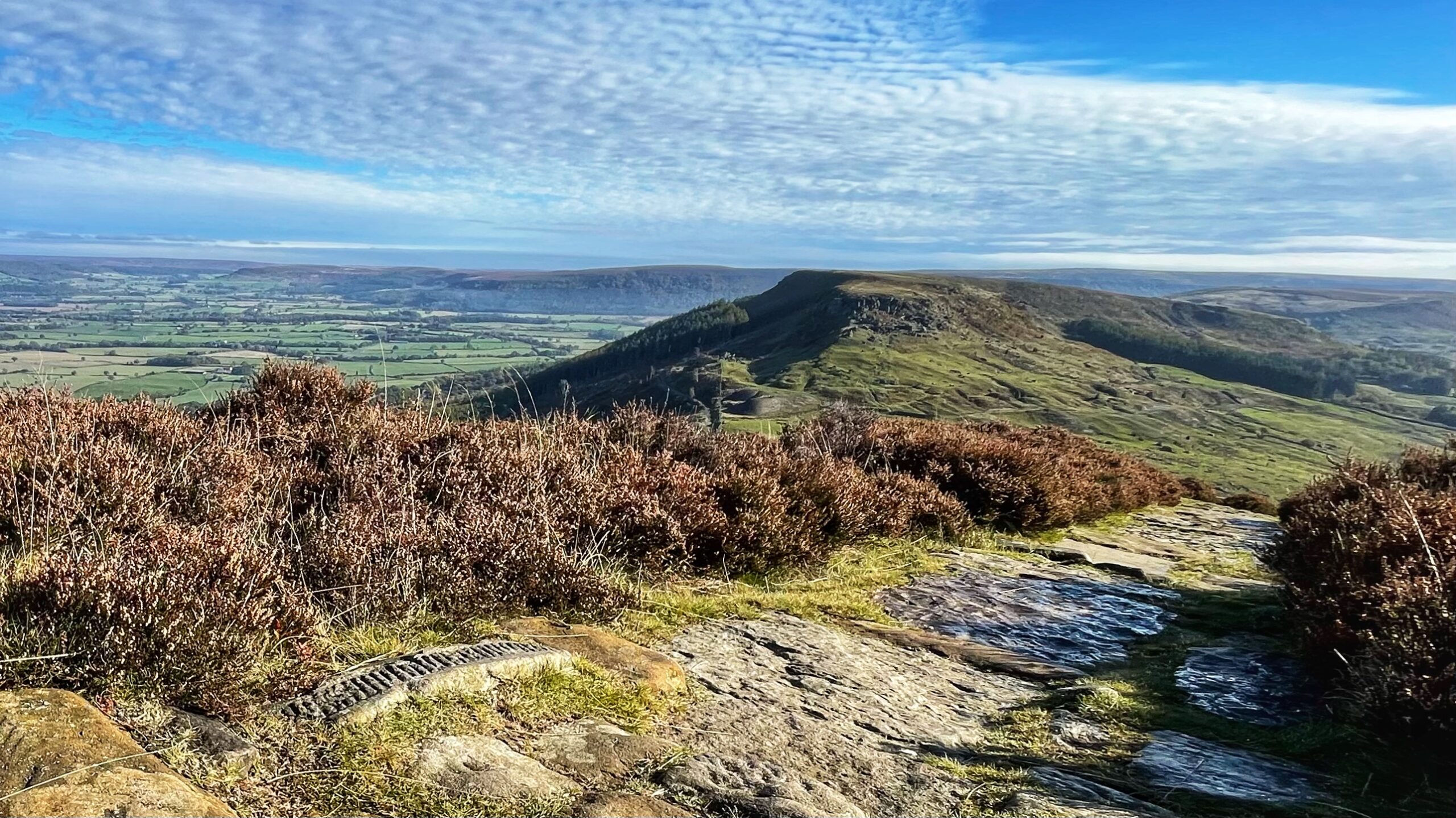 A wide-angle landscape photo of the Cleveland Hills under a bright blue sky with wispy white clouds. In the foreground, there are clumps of reddish-brown heather bordering a rocky footpath. The stone path, in the bottom left, features an distinctive slab of worked stone set flush into the ground. The path leads out toward a prominent, grass-covered hill with the distinctive crag of the Wainstones, surrounded by a vast patchwork of green fields and distant moors stretching to the horizon.