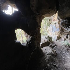 A low-light image taken from inside a small cave, looking out toward a wooded area. The interior is dark and silhouetted, dominated by the rough, textured forms of the sandstone rock ceiling and walls. There are two main openings: one to the left, which is a smaller, distinct window-like opening letting in bright light, and a larger, main exit to the right. The view through the openings shows brightly lit trees and foliage. The ground just outside the cave entrance is a hard-packed bare earth with scattered debris, and the rock wall to the right of the exit shows some colouration, possibly mineral staining.