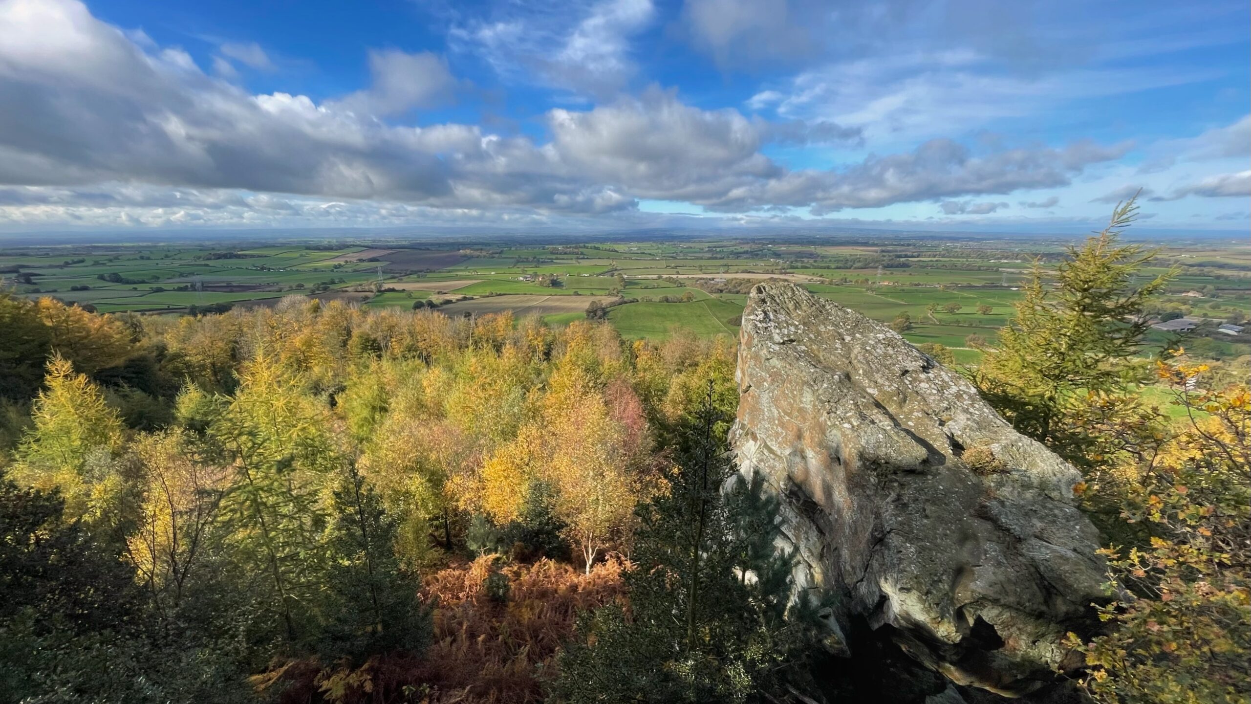 A panoramic landscape photo taken from a high vantage point, looking out over a vast, flat plain of green and brown fields. In the foreground, there's a dense wood of trees with autumnal foliage, displaying colours of yellow, gold, and green. A large, craggy rock formation dominates the right side of the foreground, partially obscuring the view. The horizon is low, and the sky is blue with scattered, dramatic white clouds.
