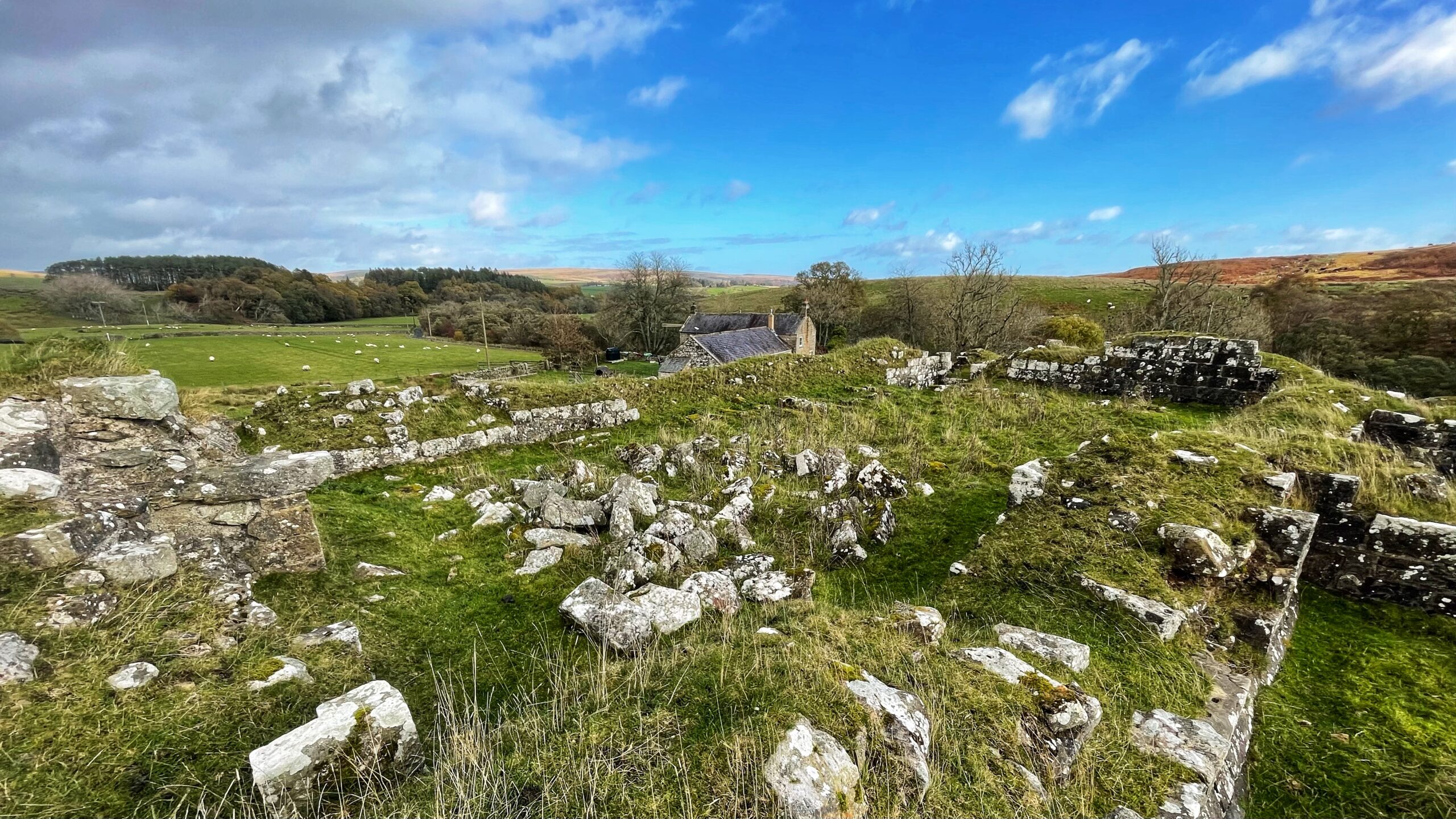 The ruined Dally Castle in Northumberland, seen from the remaining low stone foundations and rubble piles in the foreground. The foreground is a grassy mound scattered with white and grey stones, transitioning to a lush green field in the middle ground. In the distance, a small, cluster of stone buildings, Dallycastle Mill House, is visible nestled amongst trees. Rolling hills of green and brown frame the background under a bright blue sky with white, scattered clouds.