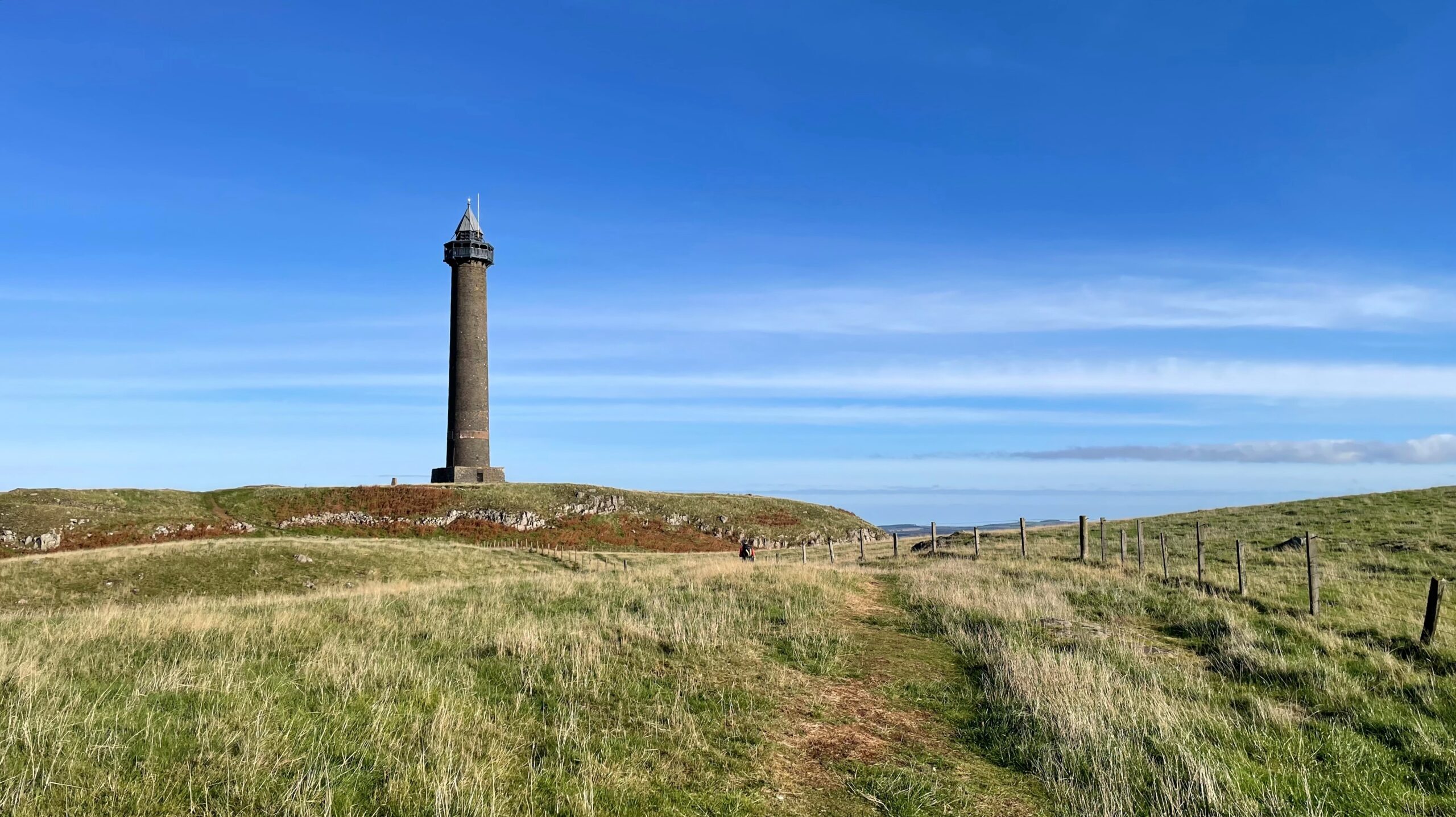 The tall, cylindrical, dark-stone Waterloo Monument stands prominently against a bright blue sky on the grassy hill of Peniel Heugh. A path leads up to the monument through the foreground, which is covered in long, dry grass. A wooden post-and-wire fence runs along the right side of the path, and a person is visible walking towards the base of the tower.