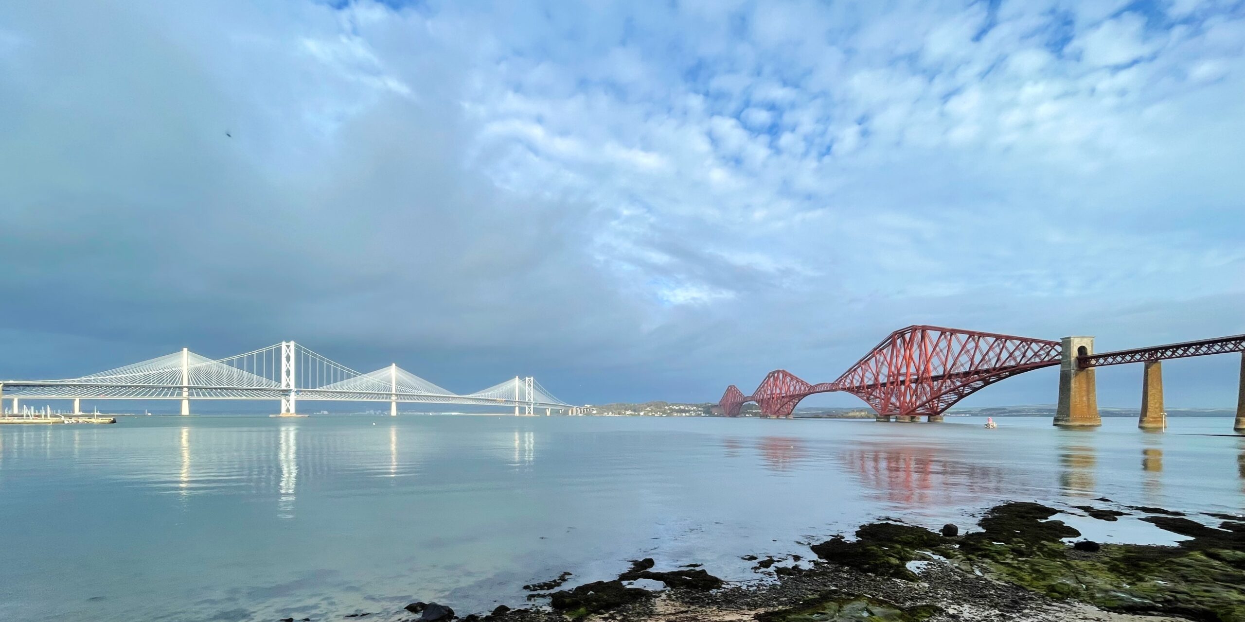 Panoramic view of the three Forth Bridges crossing the Firth of Forth in Scotland, from left to right: the white cable-stayed Queensferry Crossing road bridge behind the silver suspension Forth Road Bridge, and the iconic red cantilever Forth Bridge rail bridge.