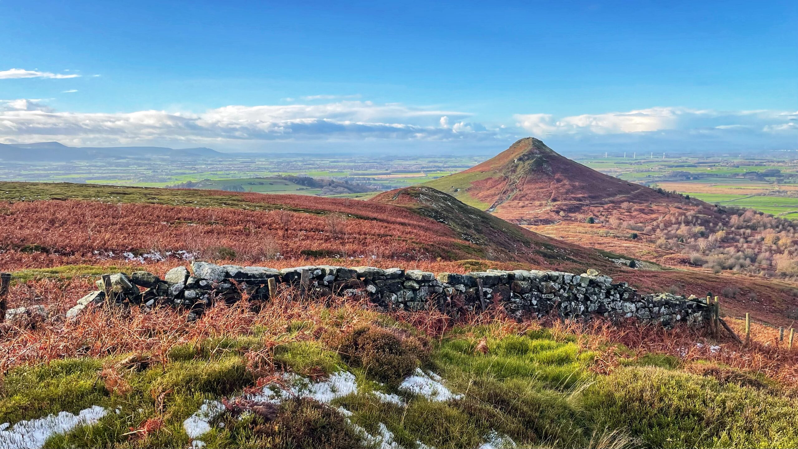 A wide-angle landscape photograph captures a crisp, clear day in North Yorkshire, dominated by the distinctive, conical peak of Roseberry Topping rising sharply in the middle-right distance, its steep slopes covered in russet-toned heather and bracken. In the foreground, a low, weathered dry stone wall stretches across the frame, dividing the immediate terrain which features patches of green and reddish-brown grasses interspersed with visible traces of light snow. Beyond the hills, the far background reveals a sprawling panorama of green fields and low, distant hills stretching toward the horizon under a bright blue sky dotted with white and grey clouds.