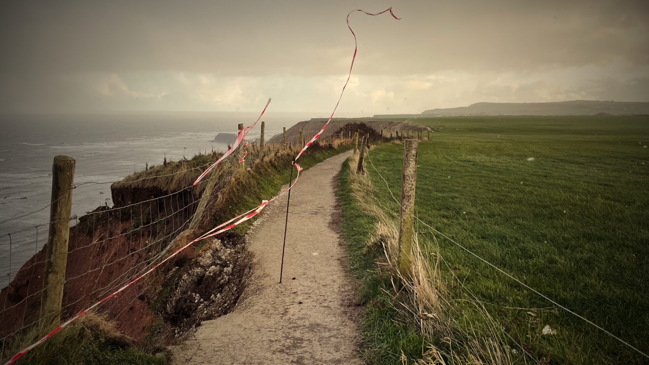 A gloomy photograph captures a dangerous section of the Cleveland Way National Trail. A narrow path is bordered on the right by a low post-and-wire fence separating it from a vast grassy field. To the left, a severe coastal cliff erosion has occurred, creating a steep, exposed drop-off of reddish-brown earth. The existing wire mesh fence along the seaward edge is significantly undermined and compromised by the cliff collapse. Red and white striped barrier tape is strung loosely between thin poles and around the void, serving as an urgent, makeshift warning sign. The ripped tape is whipping violently in the high wind, signaling the hazardous and likely closed status of the trail section due to the immediate danger of further collapse. The scene is overcast, with the rough, gray sea visible stretching out to the horizon.