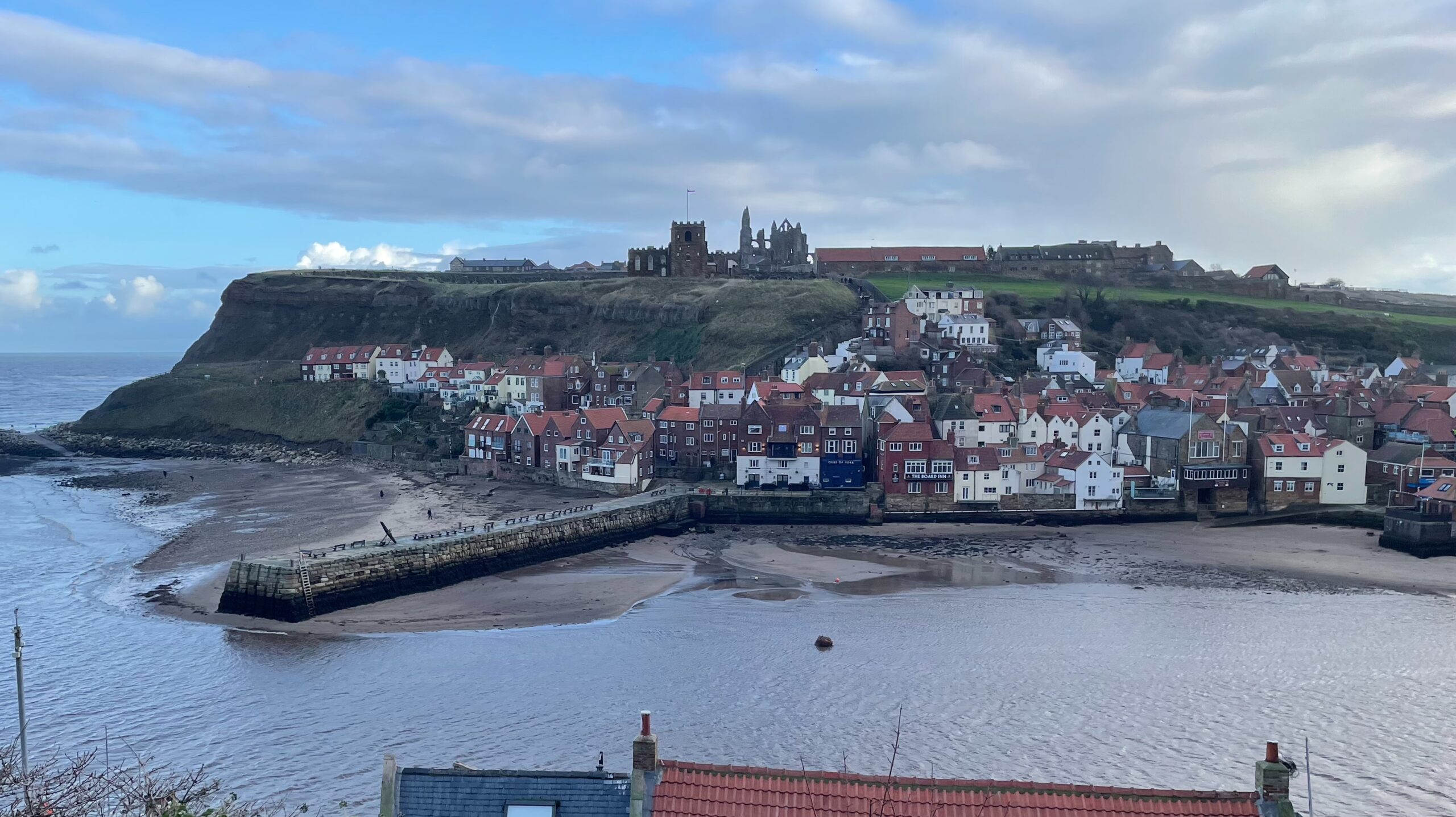 A wide-angle photo of the coastal town of Whitby. The image captures the town's lower section nestled by a sandy harbor inlet, with a long, grey stone pier extending into the water. Clustered red-roofed buildings of the town climb the steep, grassy hill in the background. Dominating the skyline atop the cliff is the ruin of Whitby Abbey and the nearby St Mary's Church, visible against a cloudy blue sky. The North Sea is visible on the left.