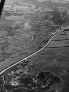 Black and white aerial photograph showing the initial construction of the Cod Beck Reservoir in the late 1940s. A long, straight road runs through the center of the frame, separating the rough, hilly terrain in the foreground from a wide, muddy valley where the dam is being built. Construction machinery is visible near a plume of steam or dust in the valley, and the surrounding land is divided into numerous agricultural fields. Distant farm buildings are scattered across the upper background.