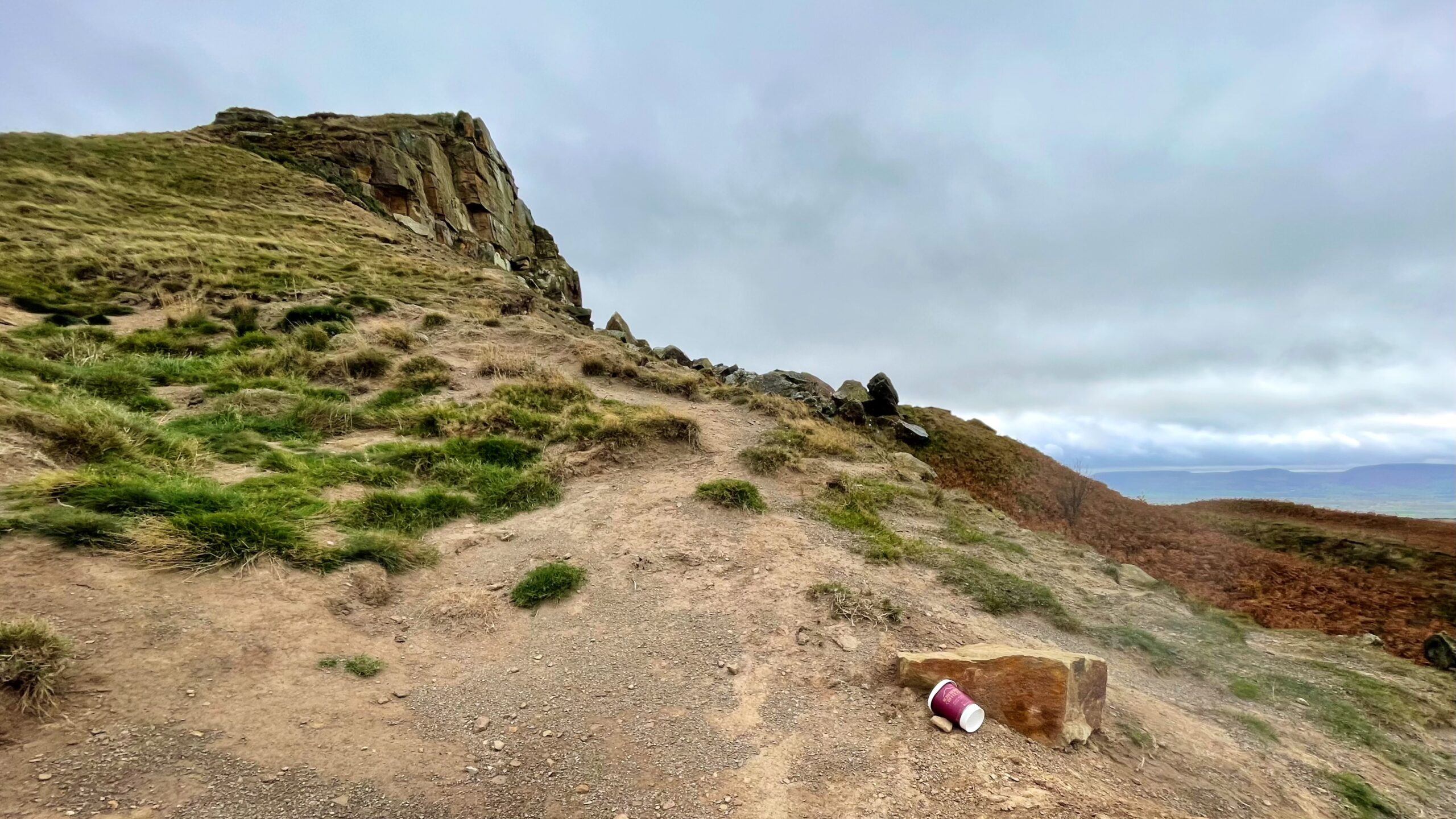 A steep, bare earth and rock-strewn slope ascending Roseberry Topping under an overcast sky. The wide, visible line of bare earth is a significant erosion scar caused by foot traffic, leading towards the rocky summit feature on the left. In the foreground, a discarded takeaway coffee cup is lying on a small, reddish-brown rock.