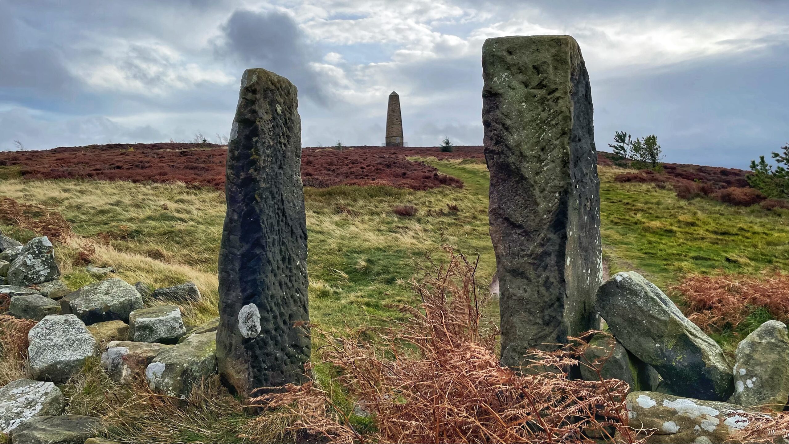 A stone monument, known as Captain Cook's Monument, stands on a grassy, heather-covered hill under a cloudy sky. In the foreground, two upright stone gateposts, heavily weathered and patterned with light-coloured lichen, frame the path leading up to the obelisk in the distance. A low stone wall with scattered boulders and bracken, also covered in lichen, is visible at the base of the gateposts.