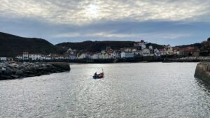 A wide shot of the harbour at Staithes, showing a cluster of traditional white and coloured cottages built into the steep hillsides. The grey water of the harbour fills the foreground, with a small, brightly coloured fishing boat near the centre. The sky is overcast but the sun is breaking through the clouds at the top.