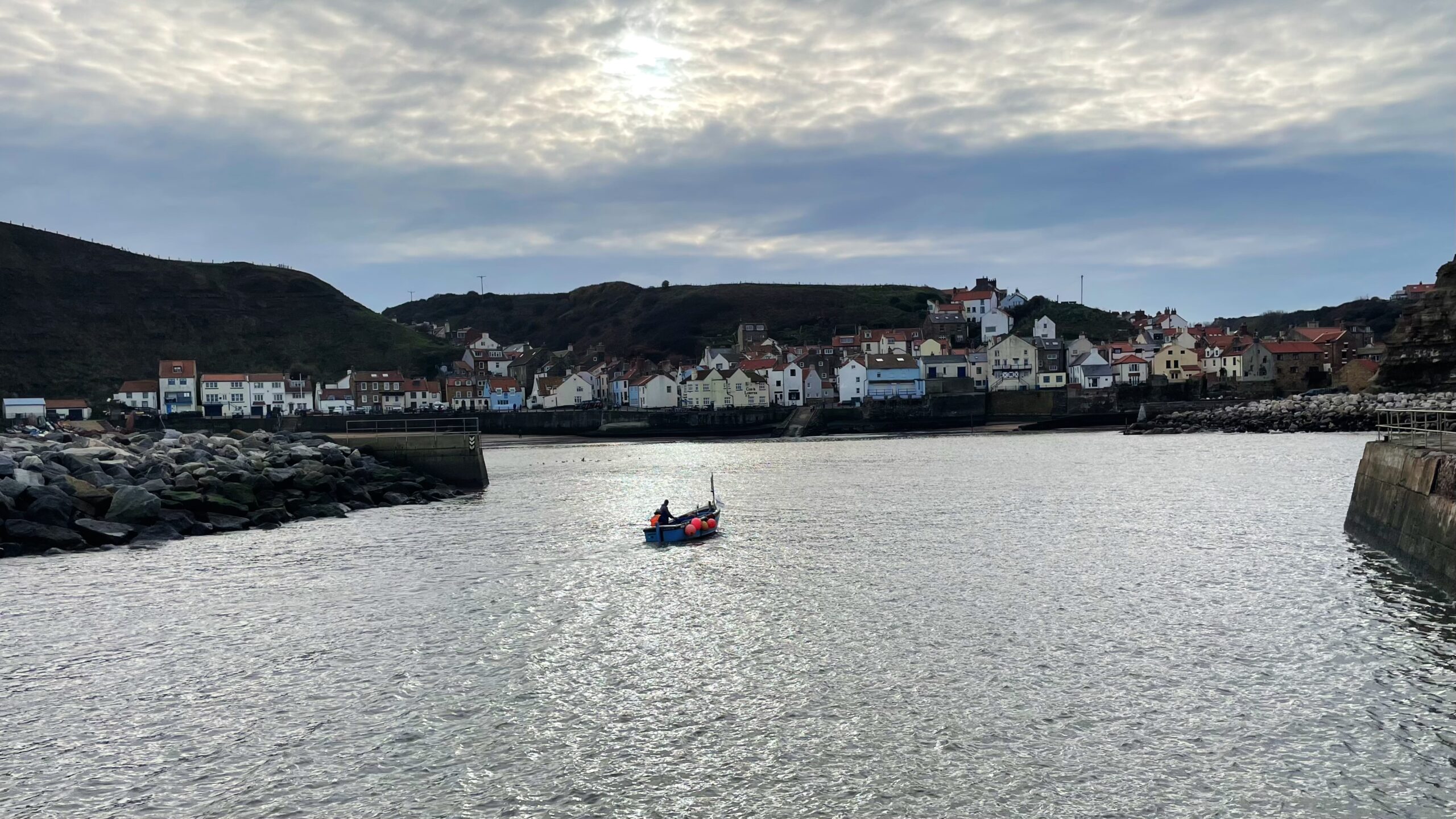 A wide shot of the harbour at Staithes, showing a cluster of traditional white and coloured cottages built into the steep hillsides. The grey water of the harbour fills the foreground, with a small, brightly coloured fishing boat near the centre. The sky is overcast but the sun is breaking through the clouds at the top.