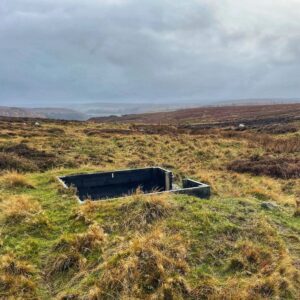 A dark, rectangular wooden shooting butt is partially sunken into a moorland landscape of tall, dry grass and heather. The moor stretches out toward a distant valley under a dramatic, cloudy grey sky. Two sheep are clearly visible grazing in the mid-ground, another three are partially obscured. The shooting butt is identified by the number ‘1’ and appears weathered. There is an entrance on the right-hand side.