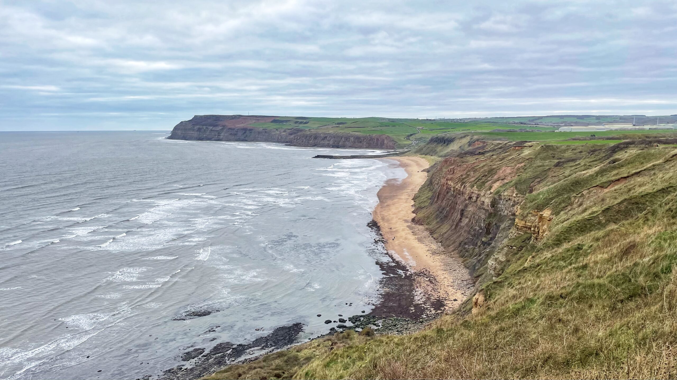 A panoramic view from a grassy cliff overlooking Cattersty Sands, a long, curving beach on the North Yorkshire coast. The sea, a grey-blue, churns with white waves and stretches out to the left. The beach is a mix of golden sand and darker rocks/shingle near the cliffs. Dramatic, stratified cliffs rise steeply on the right, covered in green and brown grasses on the upper slopes. In the distance, the cliff line culminates in a headland, beyond which are flatter green fields and some industrial structures visible on the horizon. The sky is overcast with heavy grey and white clouds.