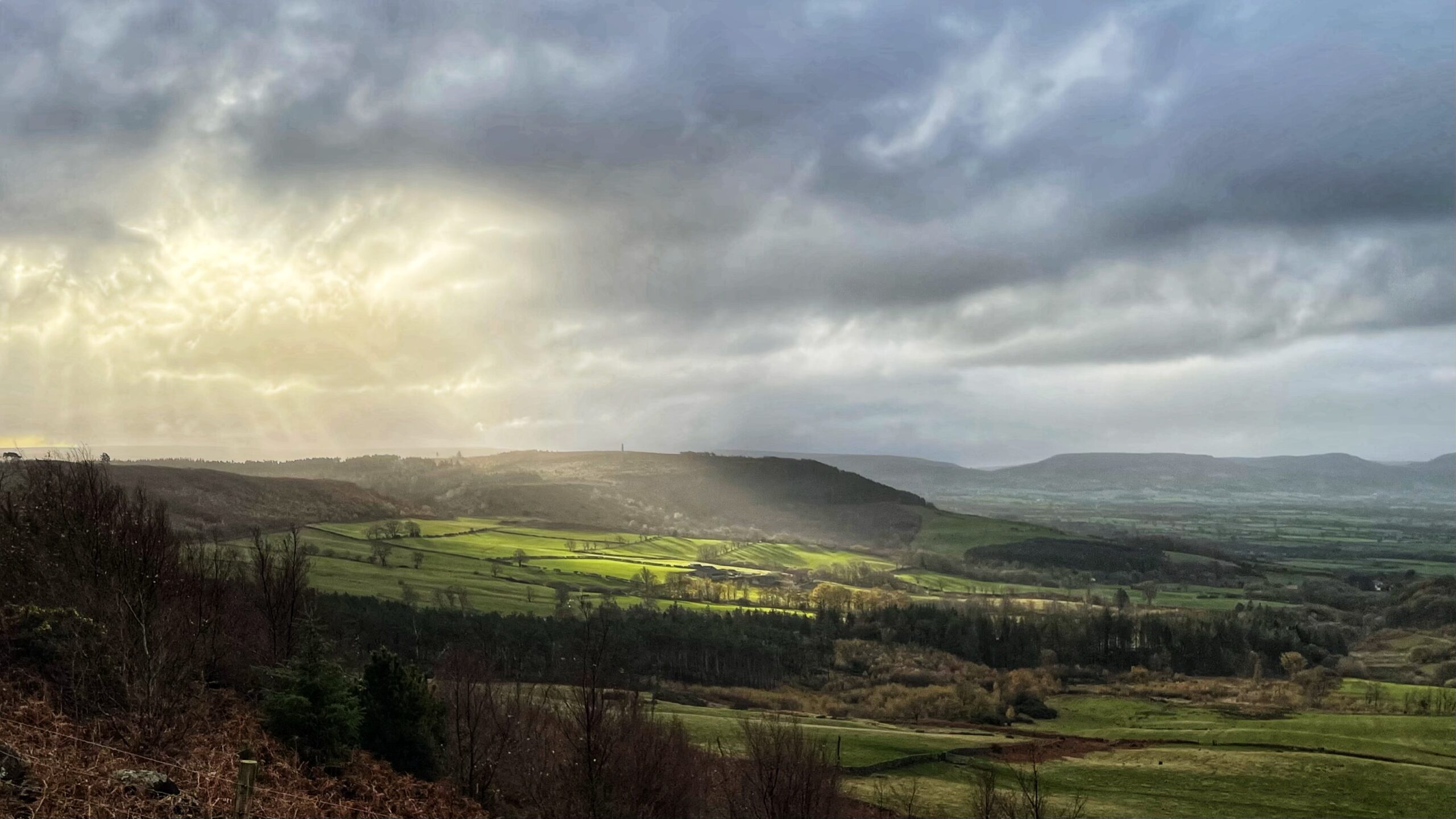 A dramatic landscape photograph under a cloudy sky. The foreground consists of dark, sparse scrubland and trees on a hillside. The middle ground is a valley of bright green fields and dark woodland. A powerful burst of sunlight, forming distinct rays or "God rays," breaks through the heavy clouds on the left and illuminates patches of the valley floor, creating a stark contrast between light and shadow. In the distance, rolling hills and fields fade into the overcast horizon.