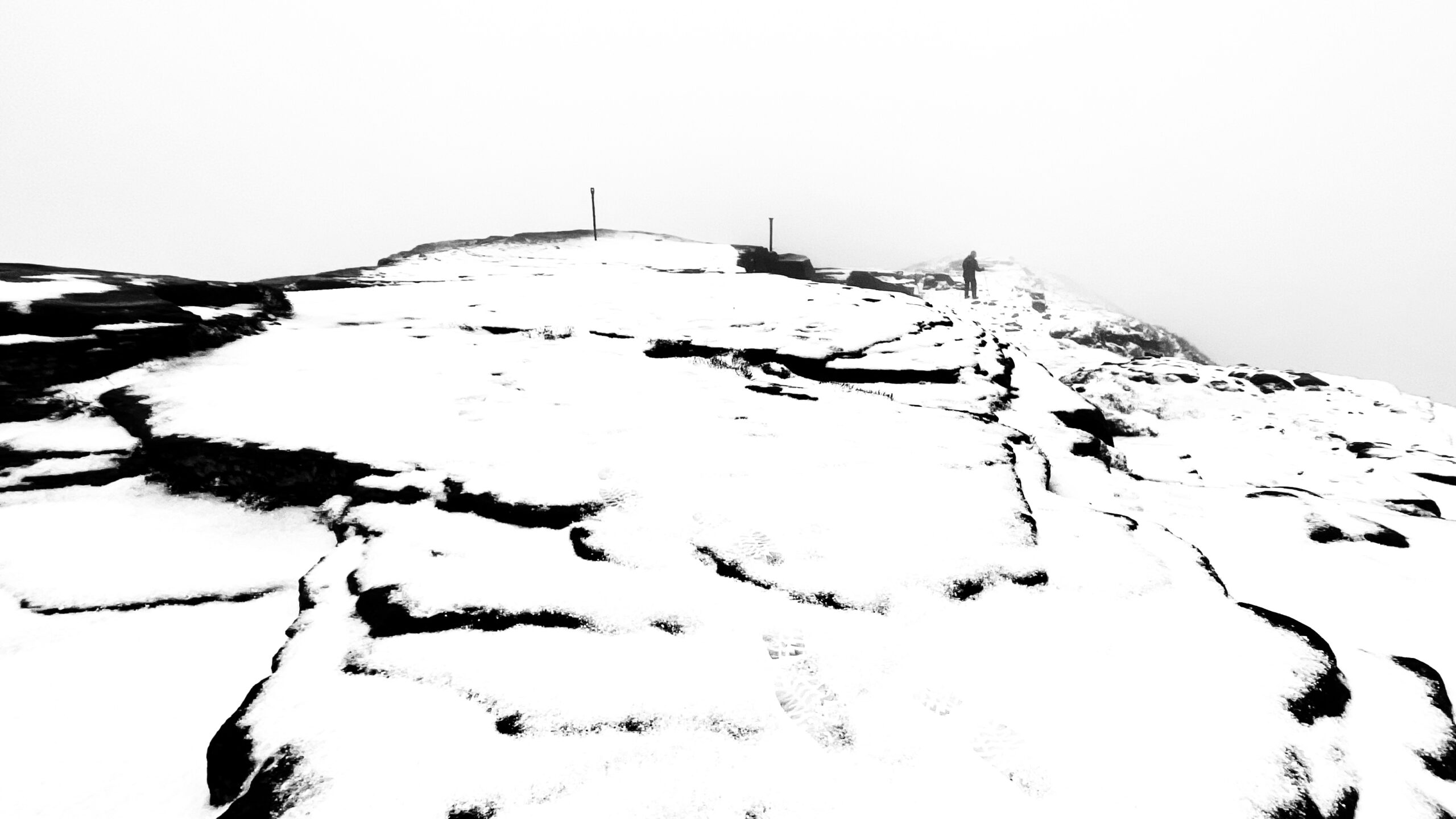 A high-contrast, black and white photograph of a snowy, rocky mountain summit shrouded in thick mist. A lone figure is visible in the distance, walking toward the peak along a ridge marked by a few vertical posts. The snow cover appears largely undisturbed, with a solitary set of footprints visible in the foreground, emphasising the desolate and challenging environment.