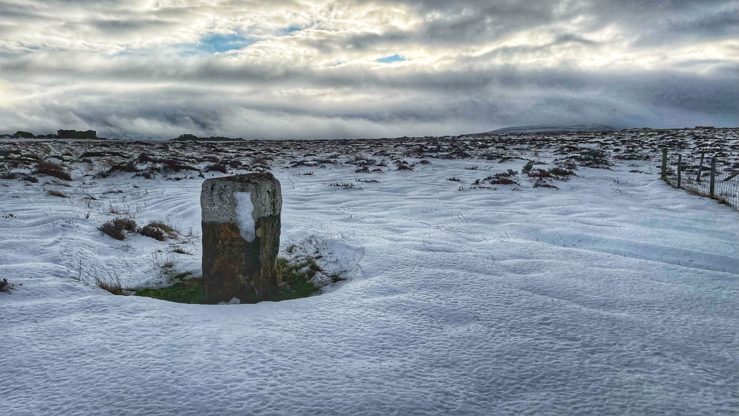A snow-covered moorland stretches under a dramatic, cloudy sky. In the foreground, a rough, squat boundary stone stands partially buried in the snow. The stone's top half shows remnants of traditional whitewash, which is significantly weathered and peeling. Low-lying brown vegetation is visible beneath the wind-blown snow, and a wire fence corner stands on the right.