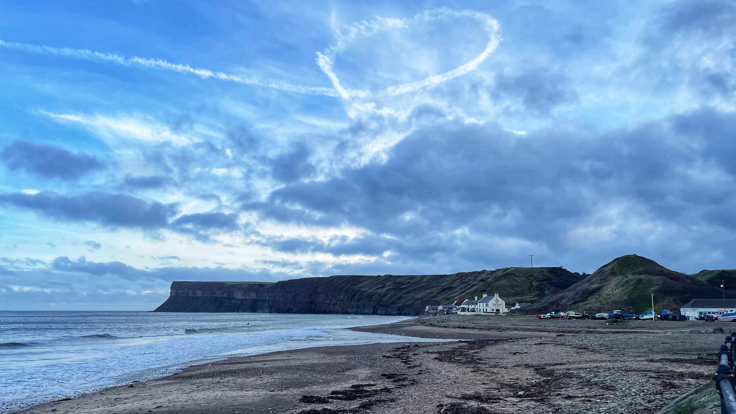 An atmospheric photograph of the coastline at Saltburn-by-the-Sea. In the foreground, a wide, dark sand and shingle beach meets the North Sea on the left. In the middle ground, a cluster of white buildings, including the Ship Inn, sits at the base of a steep grassy hill. Dominating the background is the dramatic, sheer cliff face of Huntcliff stretching horizontally into the distance. The sky is largely covered with heavy, blue-grey clouds, but a prominent white contrail forms a heart shape high above the cliffs.