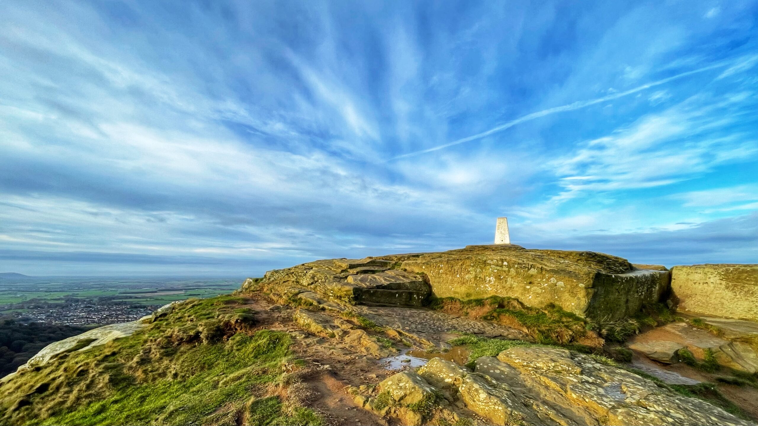 A low-angle, wide-view photo of the summit of Roseberry Topping. The triangulation pillar stands prominently in the centre-right on a rocky outcrop bathed in bright sunlight, giving the stone a warm, golden-green hue. The foreground consists of rough, mossy rocks and a grassy bank descending sharply to the left. The sky dominates the upper portion, featuring dramatic, sweeping cirrus clouds against a vibrant blue that transitions to paler tones near the horizon. In the far distance on the left, a patchwork of fields and a small settlement can be seen stretching out across the lowland plain.