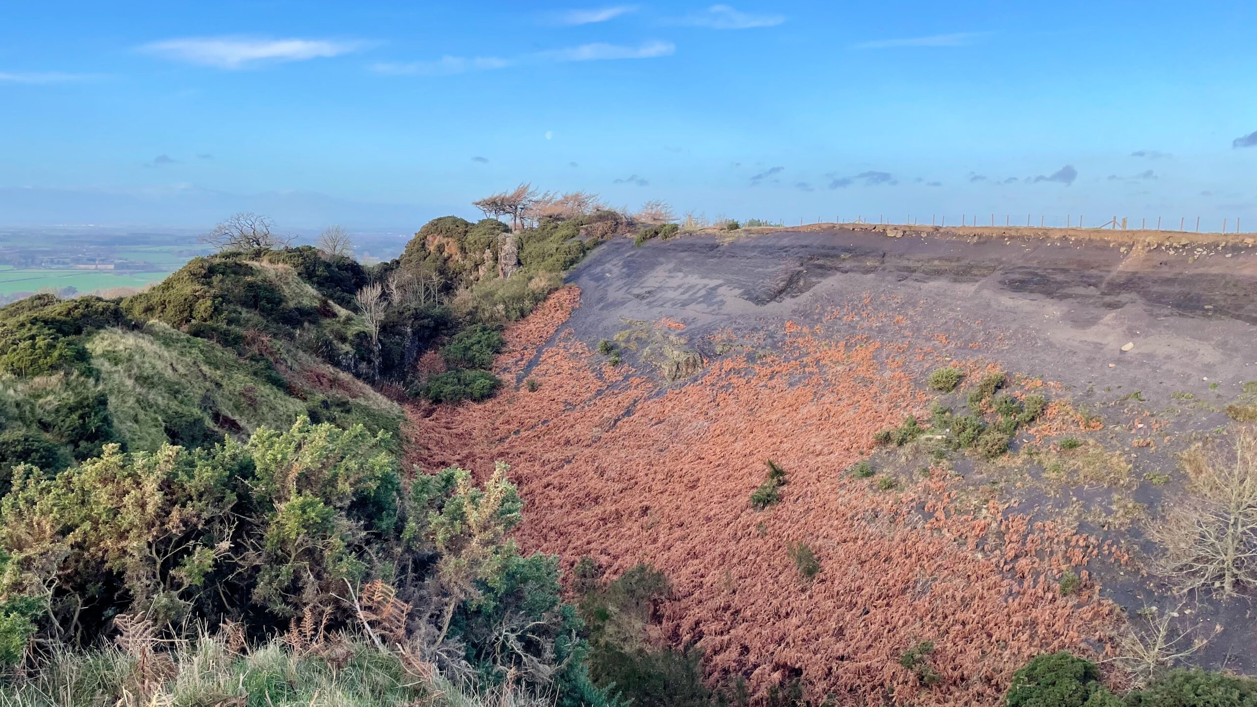 A wide, panoramic view of the abandoned Cliff Rigg quarry, showing a steep hillside. The left side of the hill is covered in green and brown scrub and gorse. The centre features a prominent, steep slope of dark shale, sparsely vegetated with rusty-red bracken and dark green shrubs. At the top right, a flat, dark area with the path along the quarry edge, extends across the top, meeting a bright blue sky with wispy clouds. The top left shows the distant, flatter landscape of the surrounding countryside.