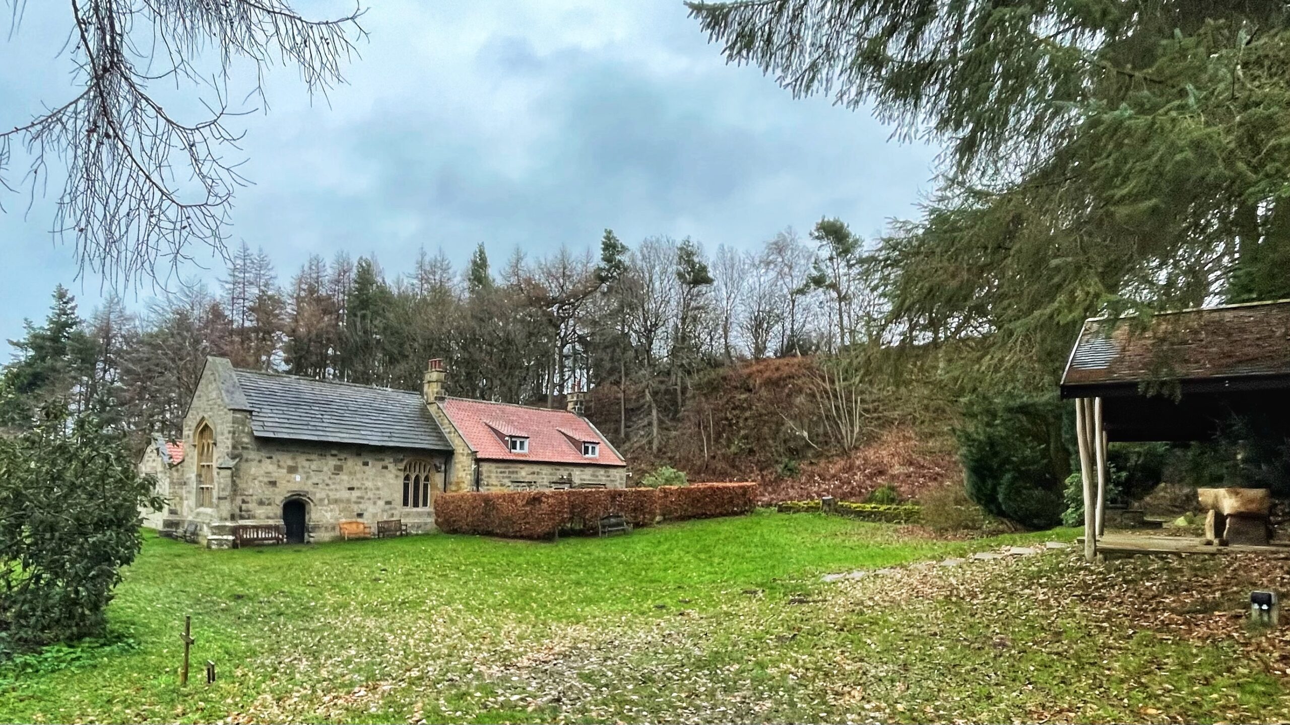 A horizontal, eye-level photo of the Lady Chapel (Shrine of Our Lady of Mount Grace) near Osmotherley, North Yorkshire, on a cloudy day. The small, stone building, which features a pointed arch window and a gabled roof with dormers on the attached cottage section, is situated in a grassy field. A low, trimmed hedge runs along the front of the cottage. To the right, a separate, open-sided wooden shelter with a dark roof stands partly visible. The background consists of a steep, wooded bank with bare and evergreen trees under a grey sky. The scene is typical of a winter or late autumn setting.