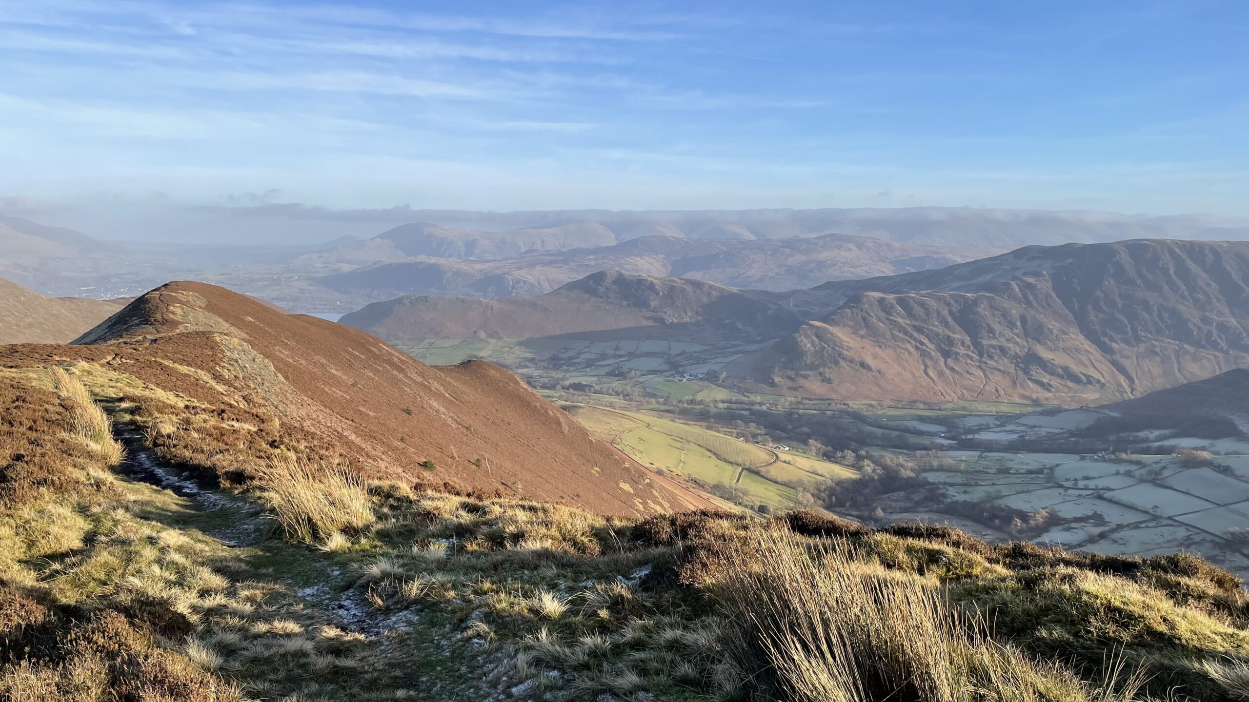 A panoramic high-angle view looking down into the Newlands Valley in the English Lake District, captured from the brown, heather-covered ridgeline of Ard Crags. In the foreground, a narrow path winds along a steep, grassy ridge that slopes down to the left. The valley floor below is a patchwork of frost-dusted green fields and stone walls, partially shaded by the surrounding fells. In the mid-ground, the distinctive rolling lower slopes of Robinson and Hindscarth rise sharply, their slopes showing a mix of deep shadows and soft morning light. The background features layers of distant, hazy blue mountains under a pale, clear sky with thin streaks of white clouds. The overall atmosphere is crisp, cold, and serene.