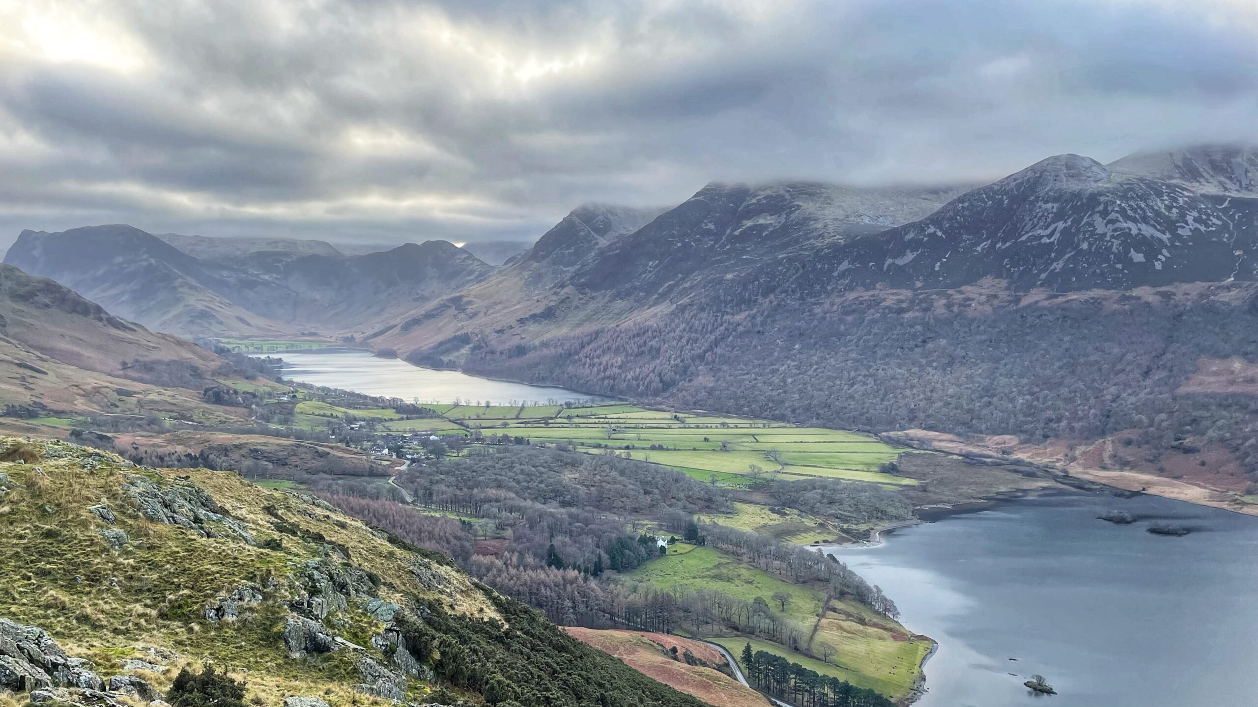 An elevated, wide-angle view of the Buttermere valley in the English Lake District under a heavy, overcast sky. In the foreground, a rocky, grass-covered hillside slopes down toward a dark, the still lake of Crummock Water on the right. A narrow strip of flat, vibrant green farmland with patchwork stone walls sits in the centre of the valley, connecting the foreground lake to a second, the lighter-coloured Buttermere lake in the distance. The valley is flanked by steep, dramatic mountains with rugged, dark grey slopes and patches of light brown vegetation. Low-hanging grey clouds partially obscure the mountain peaks and cast a soft, moody light over the entire landscape. A small cluster of white buildings and a winding road are visible nestled among the trees in the valley floor.