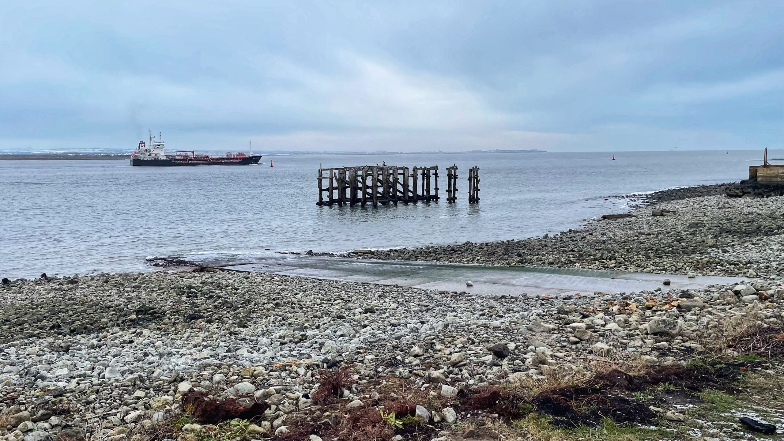 A wide-angle, eye-level shot of a rocky shoreline under a gloomy, overcast sky. In the foreground, a grey concrete slipway extends from a pebble-covered beach into the calm, dark water. Just offshore sits the skeletal remains of a weathered wooden jetty, its dark pilings standing alone in the water. To the left, a large black and red industrial tanker ship moves across the horizon. The far shoreline is visible as a thin, flat strip of land under a heavy layer of blue-grey clouds.