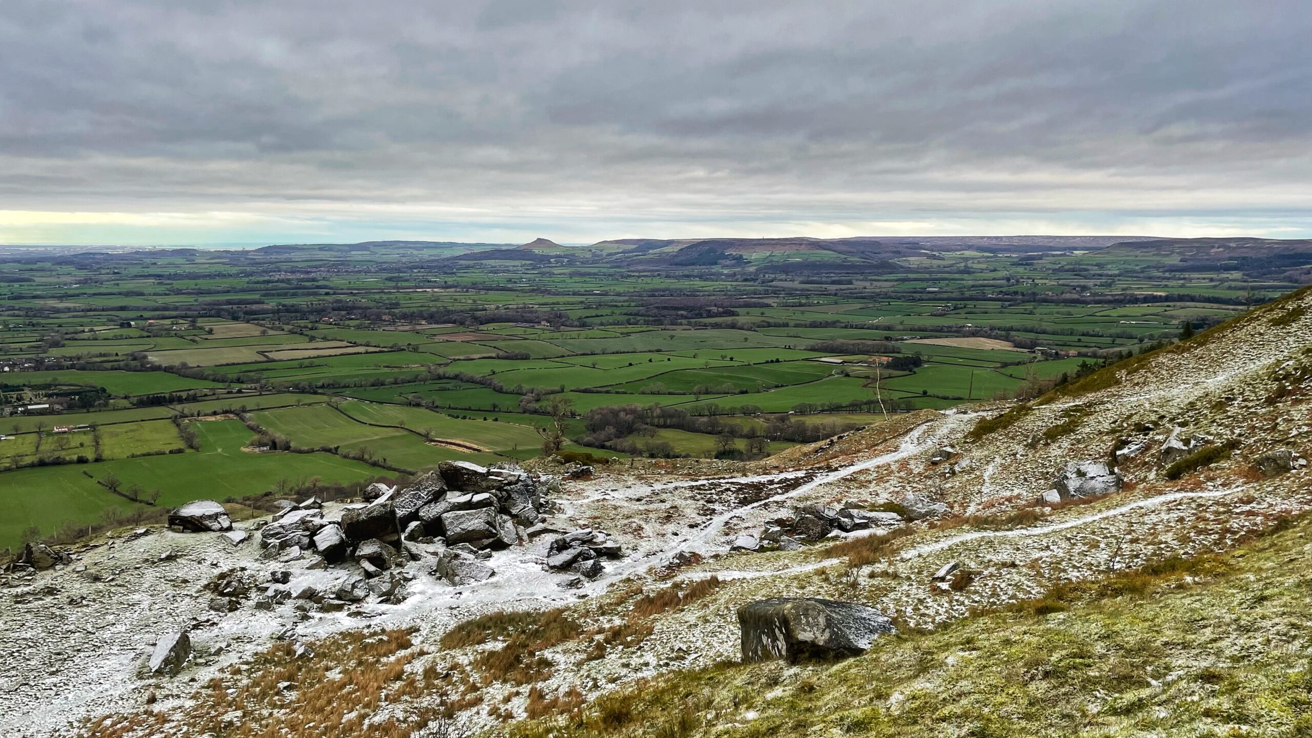 A wide-angle landscape view from a high, rocky hillside overlooking a vast valley under a cloudy, overcast sky. The foreground is a steep, grassy slope dusted with a light layer of snow, featuring scattered grey boulders and a winding path. In the middle ground, a patchwork of vibrant green fields and hedgerows stretches across the flat valley floor. In the far distance, rolling hills and the distinctive pointed hill of Roseberry Topping rise against the horizon beneath a thick layer of grey clouds.