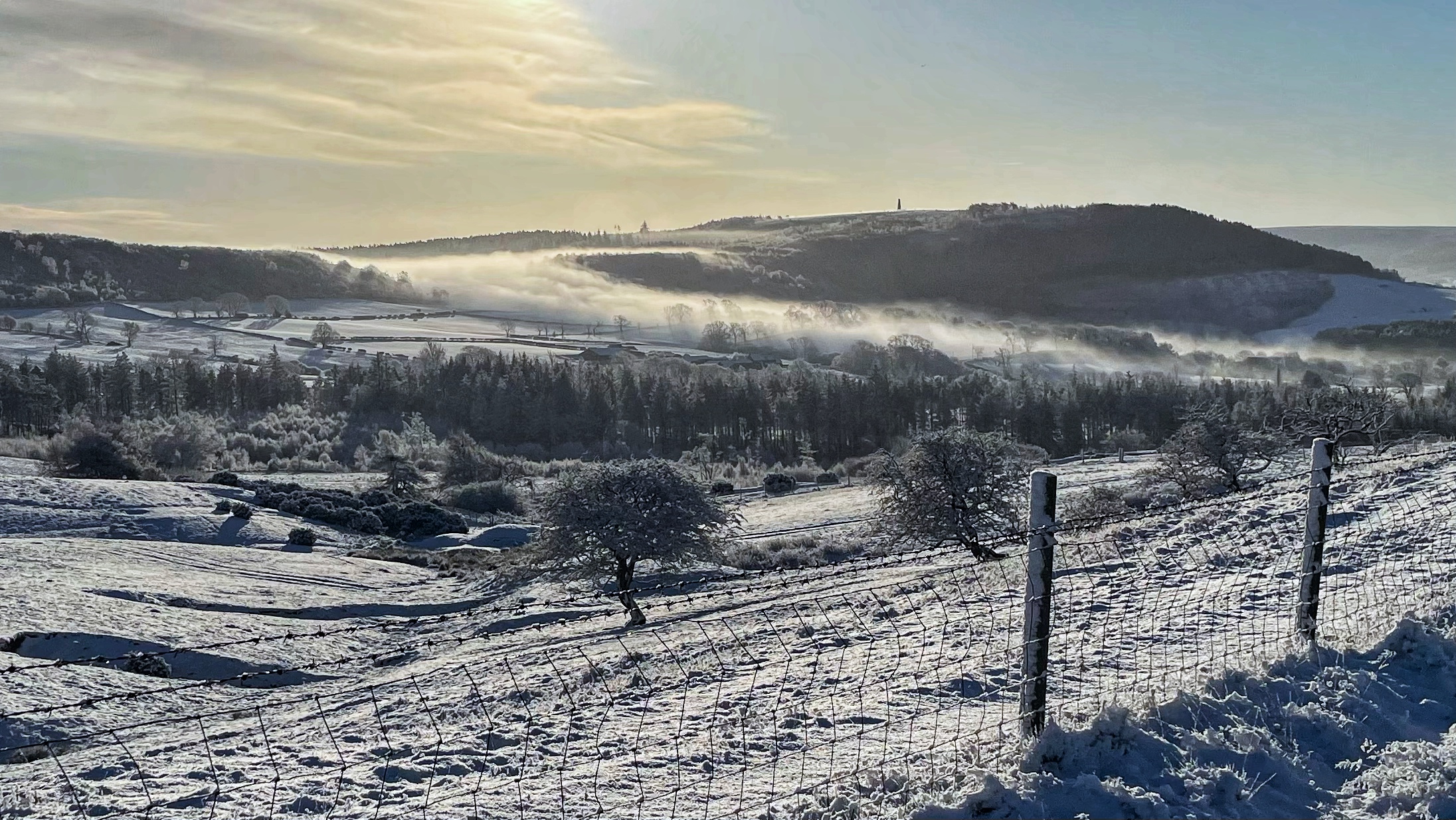 A wide-angle, eye-level landscape photograph captures a serene, snow-covered countryside under a pale, hazy winter sun. In the foreground, a wire fence with wooden posts runs diagonally from the bottom right toward the centre, its wires and mesh caked in thick white snow. The rolling hills in the mid-ground are blanketed in a layer of crisp snow, dotted with frosted trees and dark patches of evergreen forest. A dense layer of white mist or low-lying cloud settles into the valley, snaking through a col in the hills like a river. In the background, a large, dark, tree-covered ridge rises against the sky, with a tall, thin monument visible on its peak. The sky is a soft, muted blue-grey, with the sun obscured by a thin veil of clouds, casting a gentle, golden glow over the frozen scene.