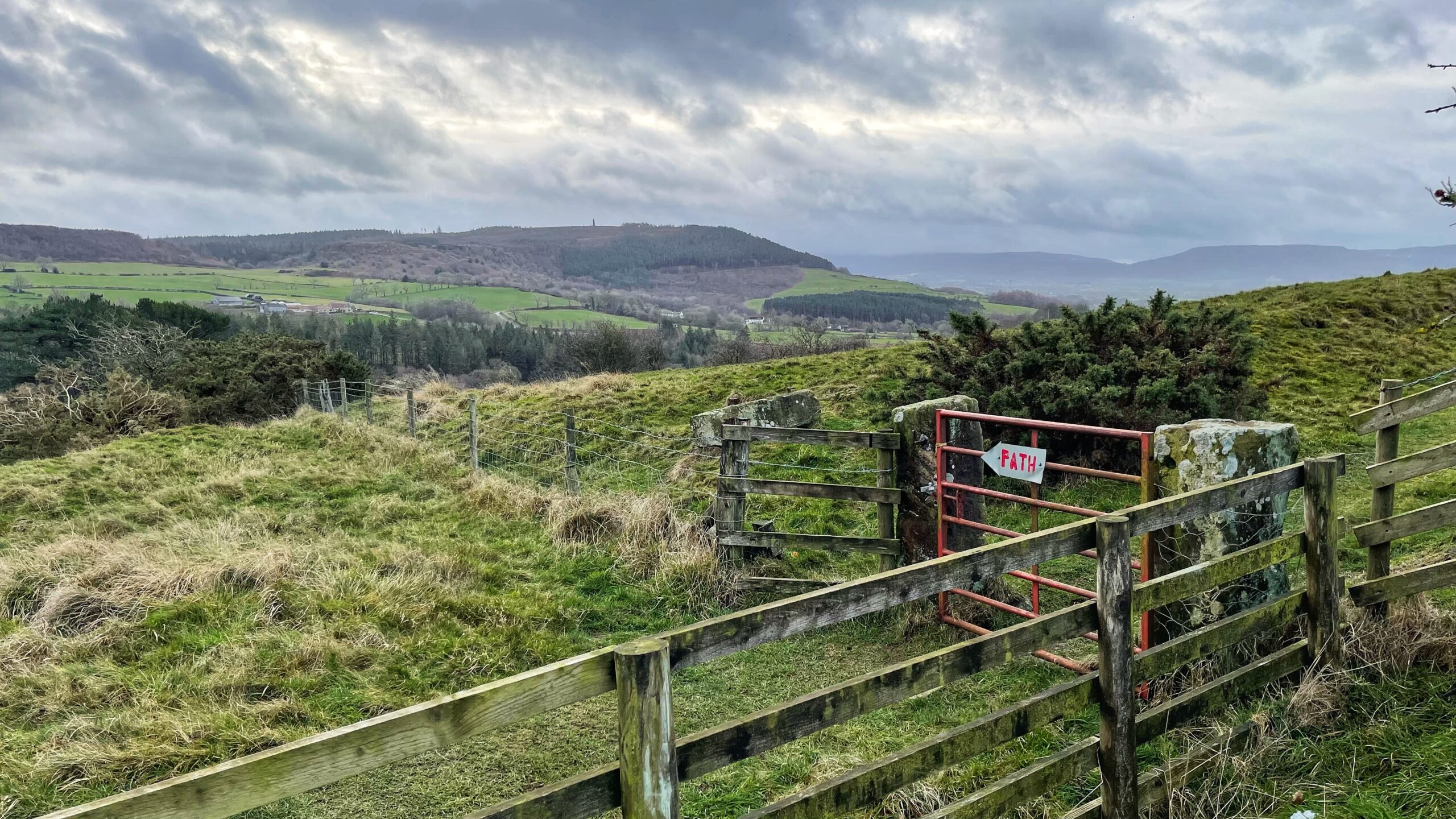 A wide landscape shot of a rolling green countryside under a cloudy, overcast sky. In the foreground, a rustic wooden and wire fence runs along a grassy hill, leading to a red metal gate. Attached to the gate is a small white sign shaped like an arrow pointing to the left, with the word "PATH" written on it in red letters. In the background, lush green fields and dense patches of forest transition into distant, hazy blue hills.