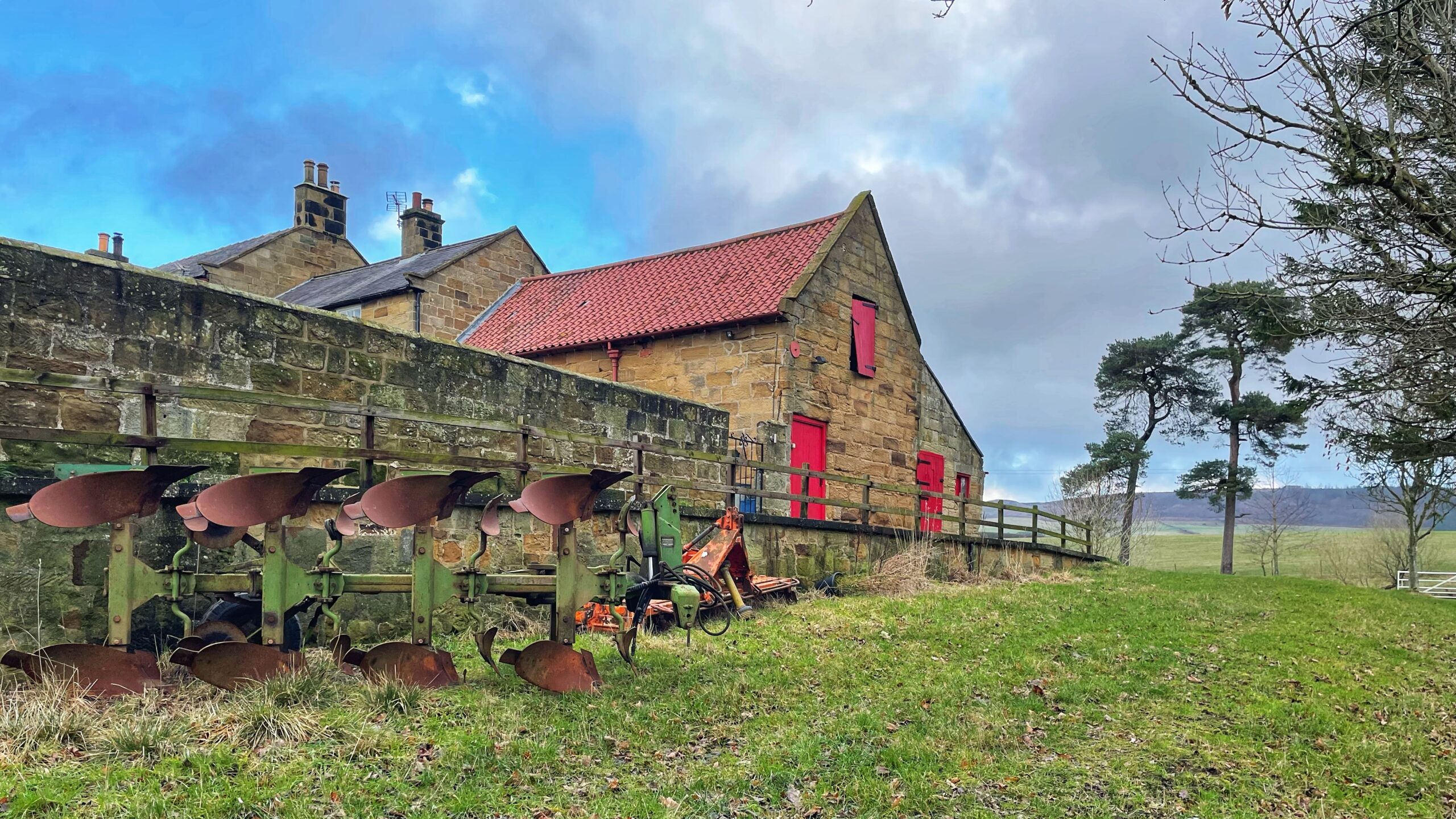 A scenic rural landscape featuring a rustic stone farm building with a red tiled roof and vibrant red doors and shutters. In the foreground, a weathered green and rusted metal plough sits on a grassy slope next to a low stone wall. In the background, there are further stone buildings with chimneys, a rolling green field, and several tall pine trees under a cloudy blue sky.