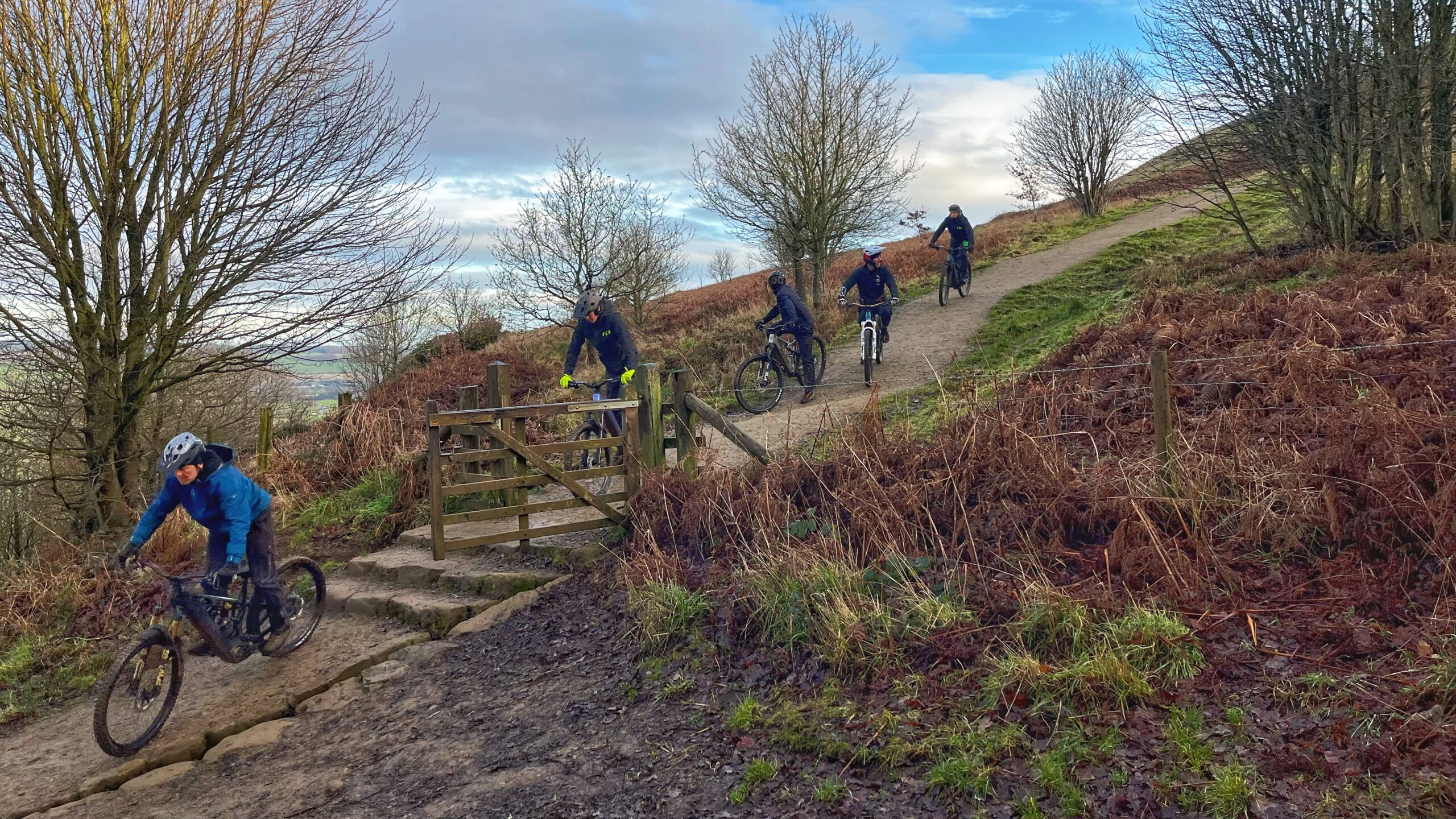 A group of five mountain bikers descends a steep, narrow, gravel path on a hillside under a cloudy sky. The lead cyclist, wearing a blue jacket and a helmet, navigates a series of stone steps. Behind them, the other four riders follow in a line down the winding path that passes through a wooden gate. The landscape is covered in brown ferns and sparse, leafless trees, suggesting a cool or wintry season.