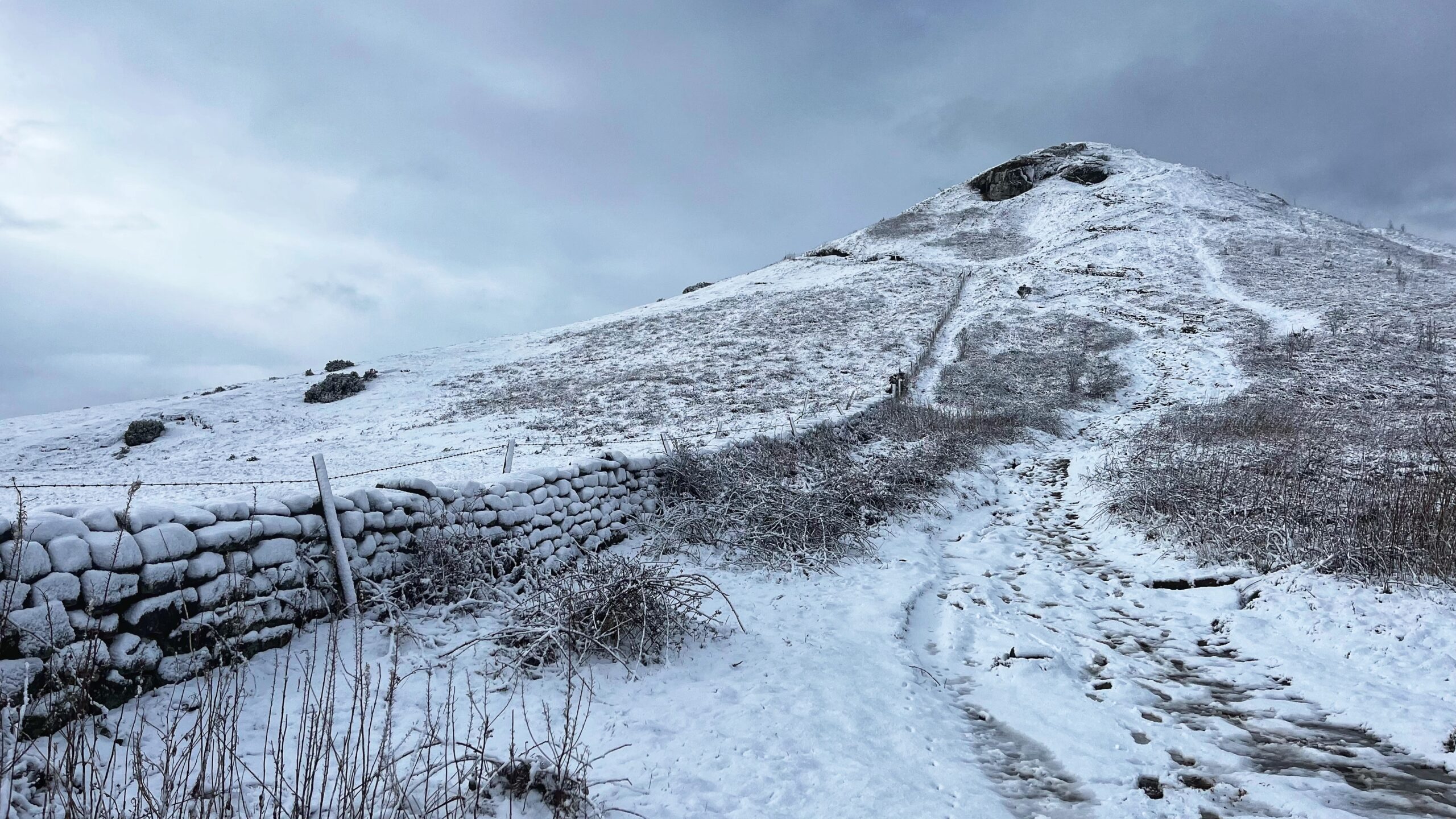 A snowy, wide-angle shot of Roseberry Topping in North Yorkshire under a heavy, overcast grey sky. In the foreground, a snow-covered stone wall runs along the left side of a slushy, footprint-laden path. The path leads the eye toward the base of the hill, where a wooden fence climbs upward. The iconic, jagged summit of the hill is draped in white, with patches of dark rock exposed near the peak. Sparse, frost-covered shrubs and dry grasses poke through the thick blanket of snow across the landscape.